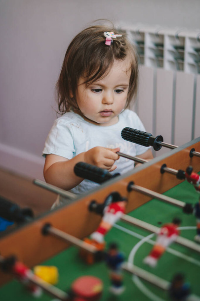 Zvláštní holčička stojící u fotbalového stolu a dívá se, jak si její rodina hraje. Šťastná rodina. Kolektivní sport. - Fotografie, Obrázek