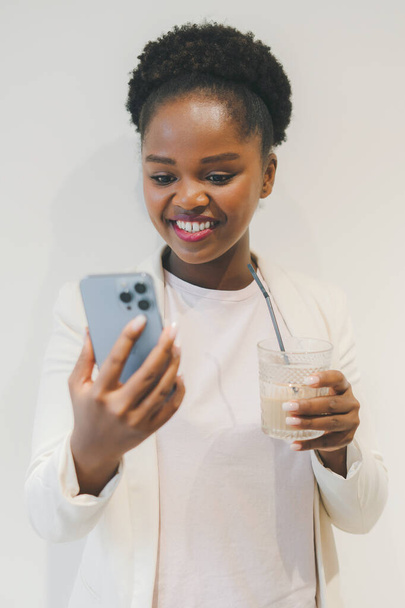 Junge hübsche Afrikanerin tippt Nachricht auf Smartphone und hält eine Kaffeetasse in der Hand, während sie isoliert über der weißen Café-Wand steht. Afrikanisches Mädchen schaut auf die - Foto, Bild