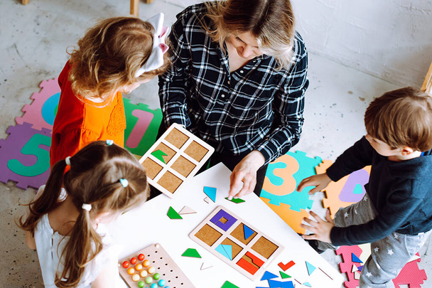 Development class for little children. Female teacher with children having fun and do smart puzzle with pieces exercises in kid development childcare center for preschoolers. High angle view - Zdjęcie, obraz