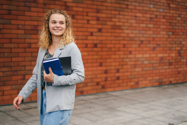 Portrét usměvavé studentky držící notebook, jak se dívá jinam a kráčí po ulici. Šťastná studentka, co jde na univerzitní kampus. Rozumím. Oběd - Fotografie, Obrázek