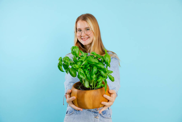Smiling cute fashionable blonde teenager girl in jeans denim presenting fresh basil plant in pot on blue isolated background. Gourmet cuisine, healthy eating, good diet and metabolism. Copy space - Photo, Image