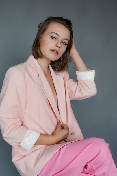 Portrait de jeune femme sexy aux yeux bleus tirant les cheveux courts et foncés, portant un pantalon rose, tenant des bords de veste, regardant la caméra, assise sur un tabouret sur fond gris. Studio, vertical. Mode. - Photo, image