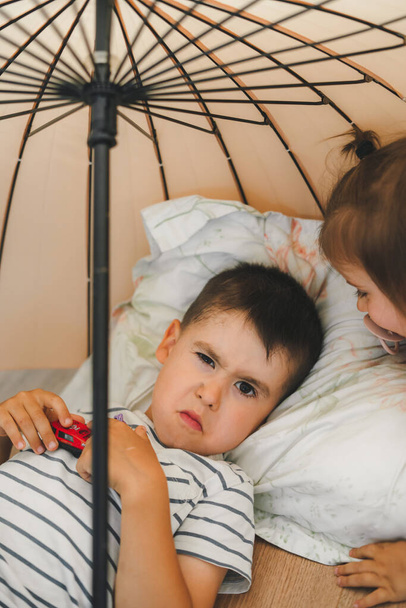 Boy glaring at the camera while lying down in the room under an umbrella next to his sister. Kids party. Fun family. - Photo, Image