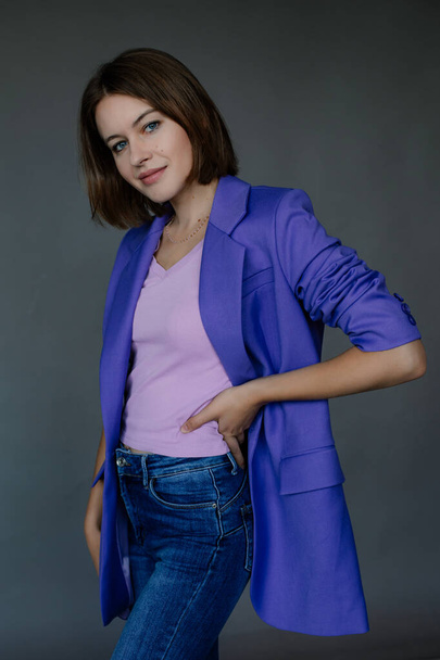 Side view of young smiling beauteous blue-eyed woman with short dark hair wearing blue jacket, jeans, looking at camera, holding hand on waist, posing on grey background. Studio, vertical. Fashion. - Foto, afbeelding