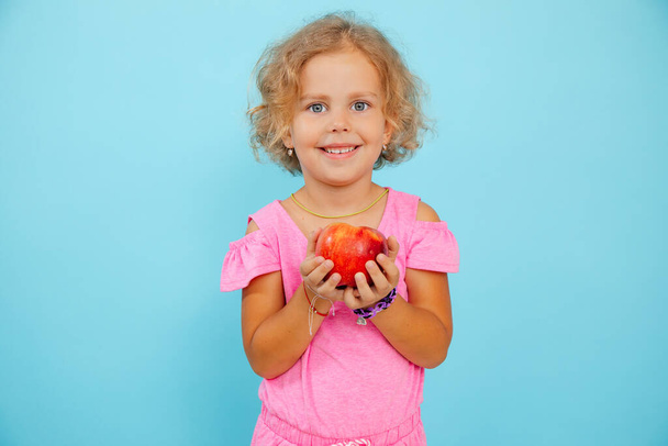 Small little smiling, expressive, positive curly blonde girl holding red juice apple in hands and looking at camera on blue isolated studio background. Healthy lifestyle for kids. Copy space - 写真・画像