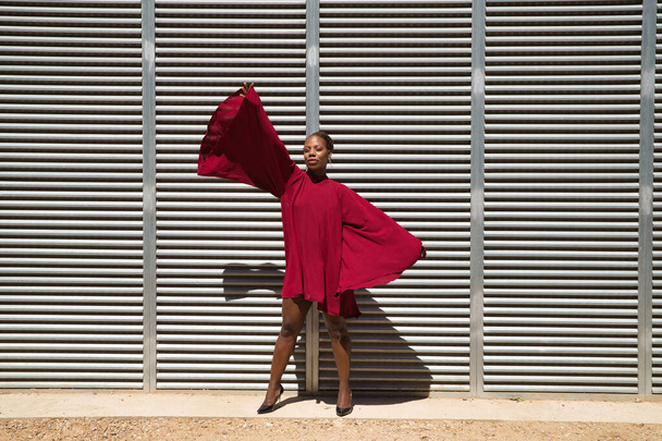 Afroameričanka v krásných červených společenských šatech s velkými rukávy pózujícími na pozadí kovových plechů. Pojem móda, krása, trend, město. - Fotografie, Obrázek