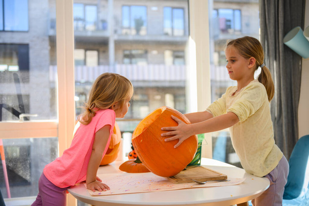 Dos chicas caucásicas lindas cortando y limpiando una calabaza, para una linterna de Halloween. Están sentados en su casa, limpiando la calabaza en la mesa.. - Foto, Imagen