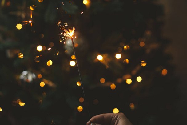 Happy New Year! Burning sparkler in female hand on background of christmas tree lights in dark room. Atmospheric celebration. Hand holding firework against stylish decorated tree with illumination - Photo, Image