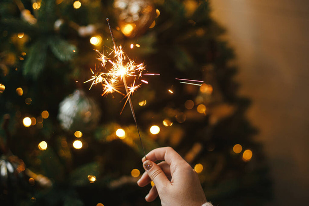 Happy New Year! Burning sparkler in female hand on background of christmas tree lights in dark room. Atmospheric celebration. Hand holding firework against stylish decorated tree with illumination - Foto, afbeelding