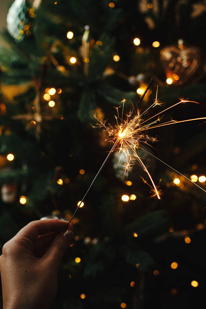 Burning sparkler in female hand on background of christmas tree lights in dark room. Happy New Year! Hand holding firework against stylish decorated tree with illumination. Atmospheric time - Φωτογραφία, εικόνα
