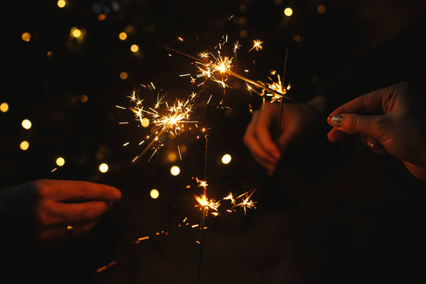 Amigos comemorando com faíscas em chamas nas mãos contra as luzes de Natal no quarto escuro. Feliz Ano Novo! Férias atmosféricas. Mãos segurando fogos de artifício no fundo da árvore iluminada elegante - Foto, Imagem