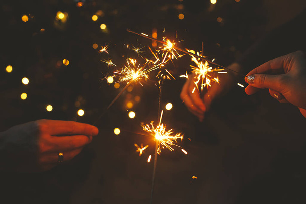 ¡Feliz Año Nuevo! Manos sosteniendo fuegos artificiales contra las luces navideñas en el cuarto oscuro. Vacaciones atmosféricas. Amigos celebrando con bengalas ardientes en las manos sobre el fondo del árbol iluminado de estilo - Foto, imagen