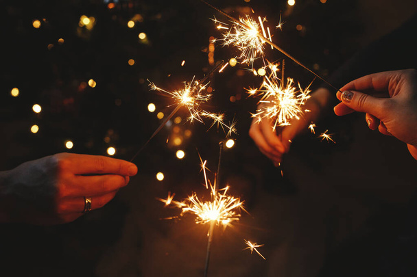 Frohes neues Jahr! Hände halten Feuerwerkskörper gegen die Weihnachtsbeleuchtung im dunklen Raum. Stimmungsvoller Urlaub. Freunde feiern mit brennenden Wunderkerzen in der Hand vor dem Hintergrund eines stilvoll beleuchteten Baumes - Foto, Bild