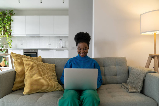 Exitosa mujer afroamericana freelancer sonriendo trabajando desde casa haciendo orden usando laptop. Chica feliz empleado de la empresa en línea hace carrera como periodista en línea o redactor se sienta en el sofá - Foto, Imagen