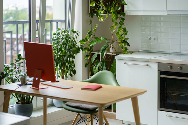 Уютное рабочее место для фрилансера со стильным красным монитором на столе рядом с большим окном и кухонной плитой. Современный домашний офис предназначен для комфортной работы для копирайтера с большим количеством комнатных растений - Фото, изображение