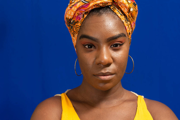 Portret afro-brazylijskiej kobiety na niebieskim tle. Nosi turban we włosach i nosi żółty.. - Zdjęcie, obraz