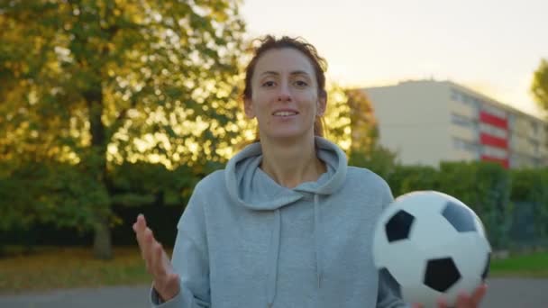 A nő, aki focilabdát tart a kamerába néz és mosolyog a napfényben. Egy női focista portréja a parkban naplementekor. Naplemente háttér. Kiváló minőségű 4k felvételek - Felvétel, videó