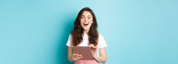 Excited smiling woman holding digital tablet, staring amazed at camera after seeing cool offer online, standing against blue background. - Foto, Imagen