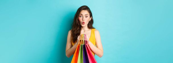 Giovane donna attraente guarda incuriosito affare promozionale, tenendo borse della spesa con merci, in piedi su sfondo blu. Copia spazio - Foto, immagini