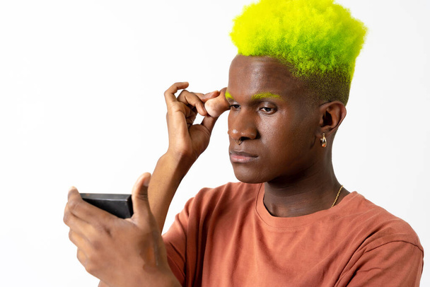 An androgynous black man posing putting on makeup, LGTBI concept, applying blush with a mirror - Фото, изображение