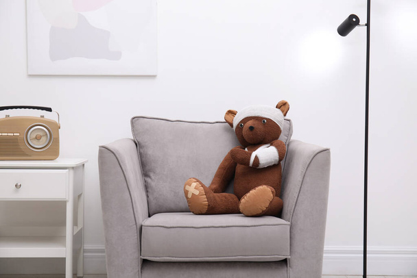 Toy bear with bandages sitting in armchair indoors - Valokuva, kuva