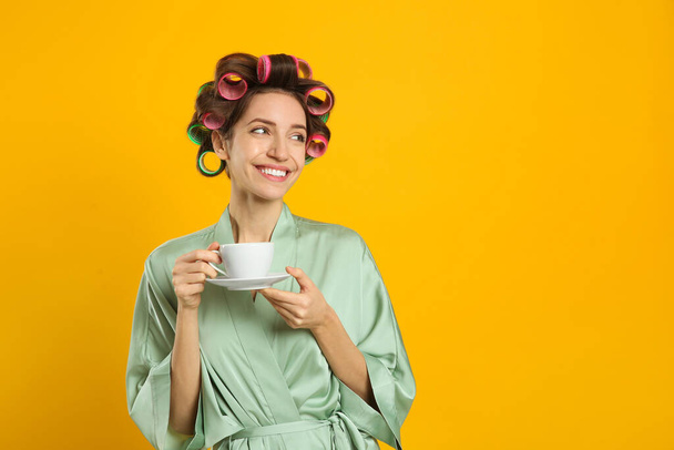 Beautiful young woman in silk bathrobe with hair curlers and cup of drink on yellow background. Space for text - Fotoğraf, Görsel