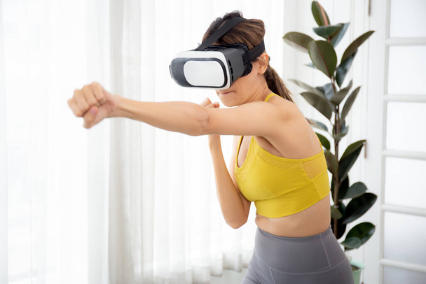 Young caucasian woman wearing vr headset while workout and exercise together at living room, futuristic and innovation of virtual reality metaverse, female playing game and punch with enjoy. - Фото, изображение
