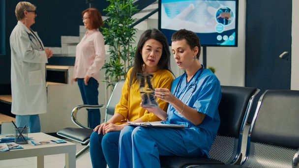 Health specialist showing radiography scan diagnosis to patient in waiting room, looking at x ray exam results with nurse in facility lobby. Analyzing orthopedic disease at medical consultation. - Foto, Imagen