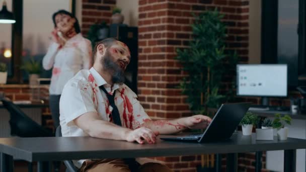 Scary looking zombie trying to work on modern laptop in office workspace. Spooky doomsday brain dead monster with deep and bloody wounds and scars trying to use portable computer in agency space. - Metraje, vídeo