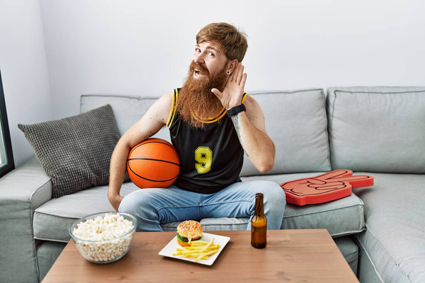 Blanke man met een lange baard die basketbalbal vasthoudt en tv-spel lacht met hand in hand luisterend naar geruchten of roddels. doofheid.  - Foto, afbeelding