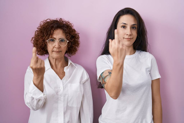 Hispanic mother and daughter wearing casual white t shirt over pink background showing middle finger, impolite and rude fuck off expression  - Zdjęcie, obraz