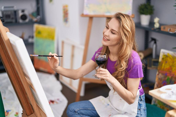 Νεαρή ξανθιά καλλιτέχνης γυναίκα πίνοντας κρασί σχέδιο στο στούντιο τέχνης - Φωτογραφία, εικόνα