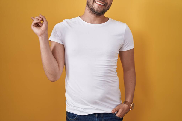 Homme hispanique portant t-shirt blanc sur fond jaune souriant heureux pointant avec la main et le doigt sur le côté  - Photo, image