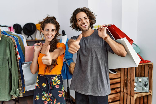 Ein junges hispanisches Paar, das im Einzelhandel Einkaufstüten in der Hand hält, zustimmend positive Gesten mit der Hand macht, lächelnd die Daumen nach oben streckt und sich über den Erfolg freut. Siegergeste.  - Foto, Bild