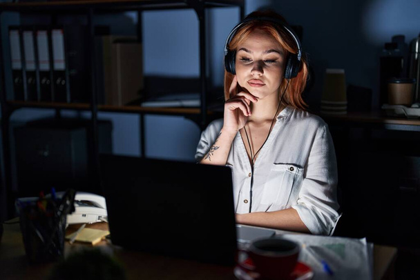 Young caucasian woman working at the office at night with hand on chin thinking about question, pensive expression. smiling with thoughtful face. doubt concept.  - 写真・画像