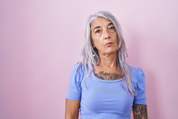 Жінка середнього віку з татуюваннями, що стоять над рожевим тлом, виглядає сонною і втомленою, виснаженою втомою і похміллям, лінивими очима вранці
.  - Фото, зображення