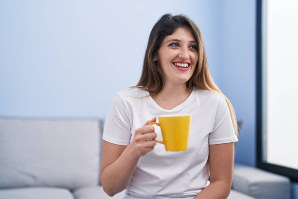 Young hispanic woman drinking coffee sitting on sofa at home - Zdjęcie, obraz