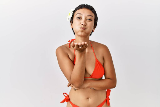 Mujer hispana joven con el pelo corto con bikini mirando a la cámara soplando un beso con la mano en el aire siendo encantadora y sexy. expresión de amor.  - Foto, imagen