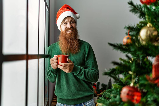Young redhead man drinking coffee standing by christmas tree at home - Fotoğraf, Görsel
