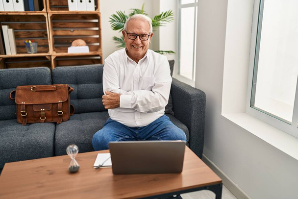 Senior therapist with grey hair doing online session at consultation office happy face smiling with crossed arms looking at the camera. positive person.  - Photo, Image