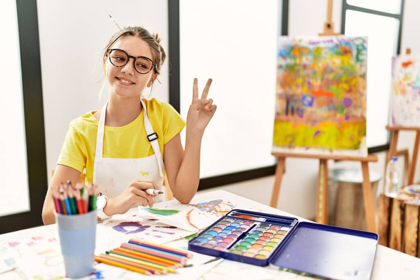 Jonge brunette tiener in de kunststudio glimlachend naar de camera kijkend met vingers die een overwinningsbord doen. nummer twee.  - Foto, afbeelding