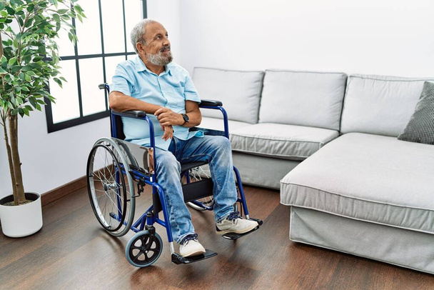 Pohledný starší muž sedící na vozíčku v obývacím pokoji a dívající se stranou s úsměvem na tváři, přirozený výraz. jistota smíchu.  - Fotografie, Obrázek