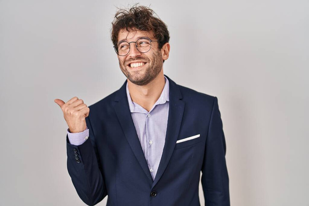 Hispanic business man wearing glasses smiling with happy face looking and pointing to the side with thumb up.  - Photo, Image