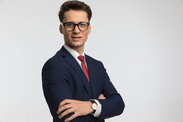 happy young businessman wearing navy blue suit crossing arms and smiling in front of grey background in studio - Fotografie, Obrázek
