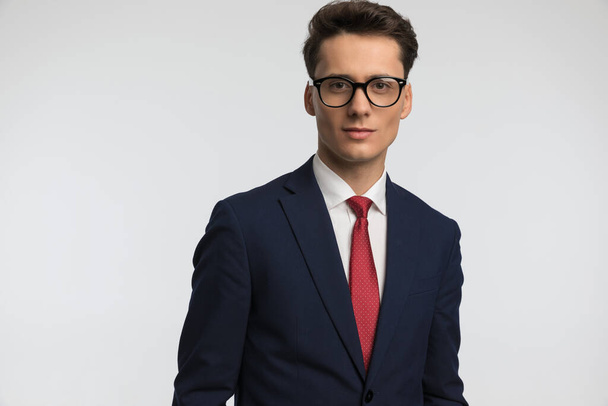 portrait of happy man with eyeglasses wearing suit with red tie and smiling in front of grey background in studio - Photo, Image