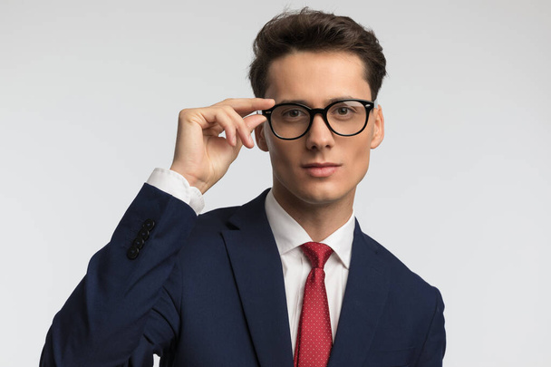 elegant businessman with red tie smiling and adjusting eyeglasses in front of grey background in studio - Foto, immagini