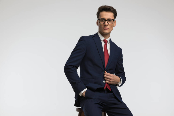 confident businessman with glasses closing suit and posing with hand in pocket while sitting in front of grey background  - Fotó, kép