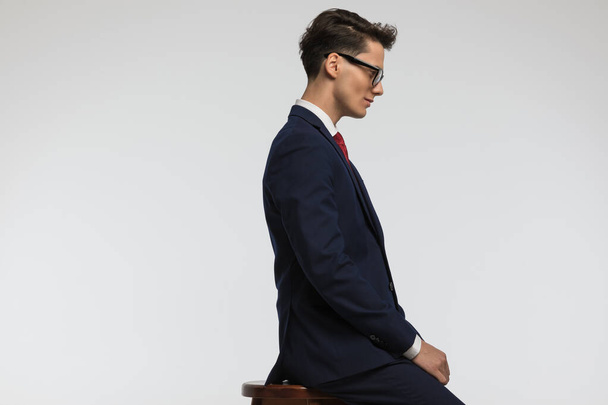 вид сидящего бизнесмена в очках, улыбающегося и глядящего вниз сбоку на фоне серого фона в студии - Фото, изображение