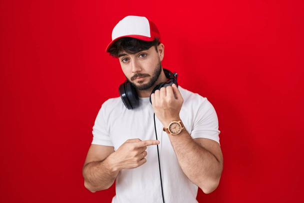 Homme hispanique avec barbe portant chapeau de joueur et écouteurs pressés pointant vers regarder le temps, l'impatience, en regardant la caméra avec une expression détendue  - Photo, image