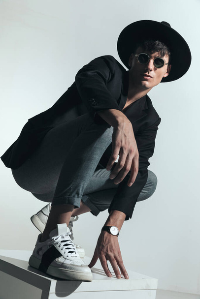 confiante jovem com chapéu preto e casaco segurando cotovelo no joelho e agachado na frente de fundo cinza no estúdio, sendo legal e posando de uma forma - Foto, Imagem
